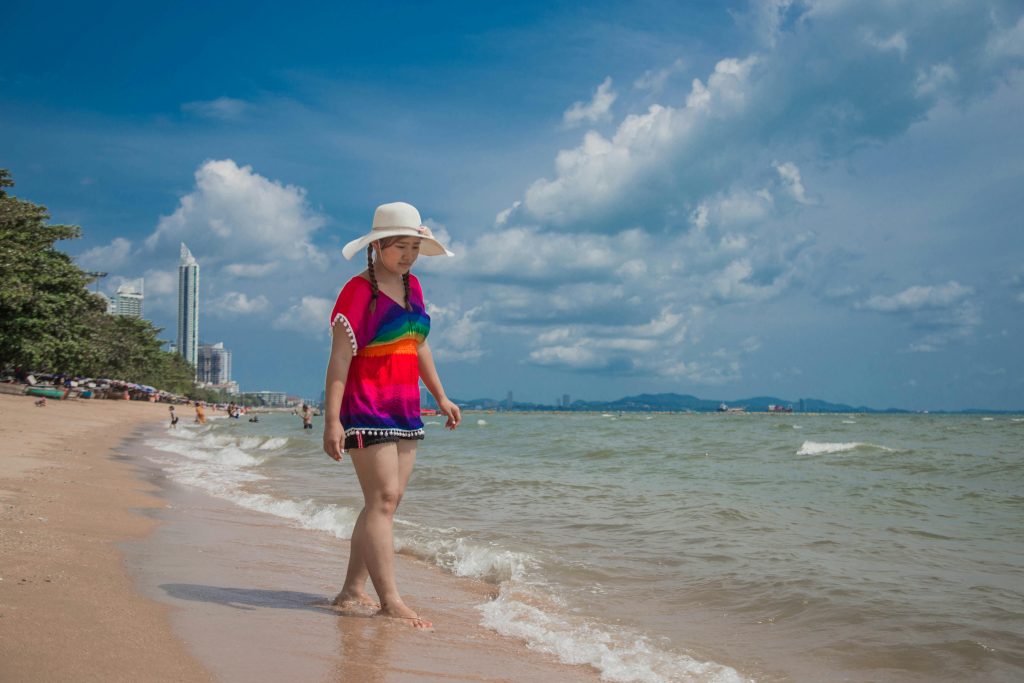 A young woman in a hat walks barefoot along a tropical beach under a bright sky.